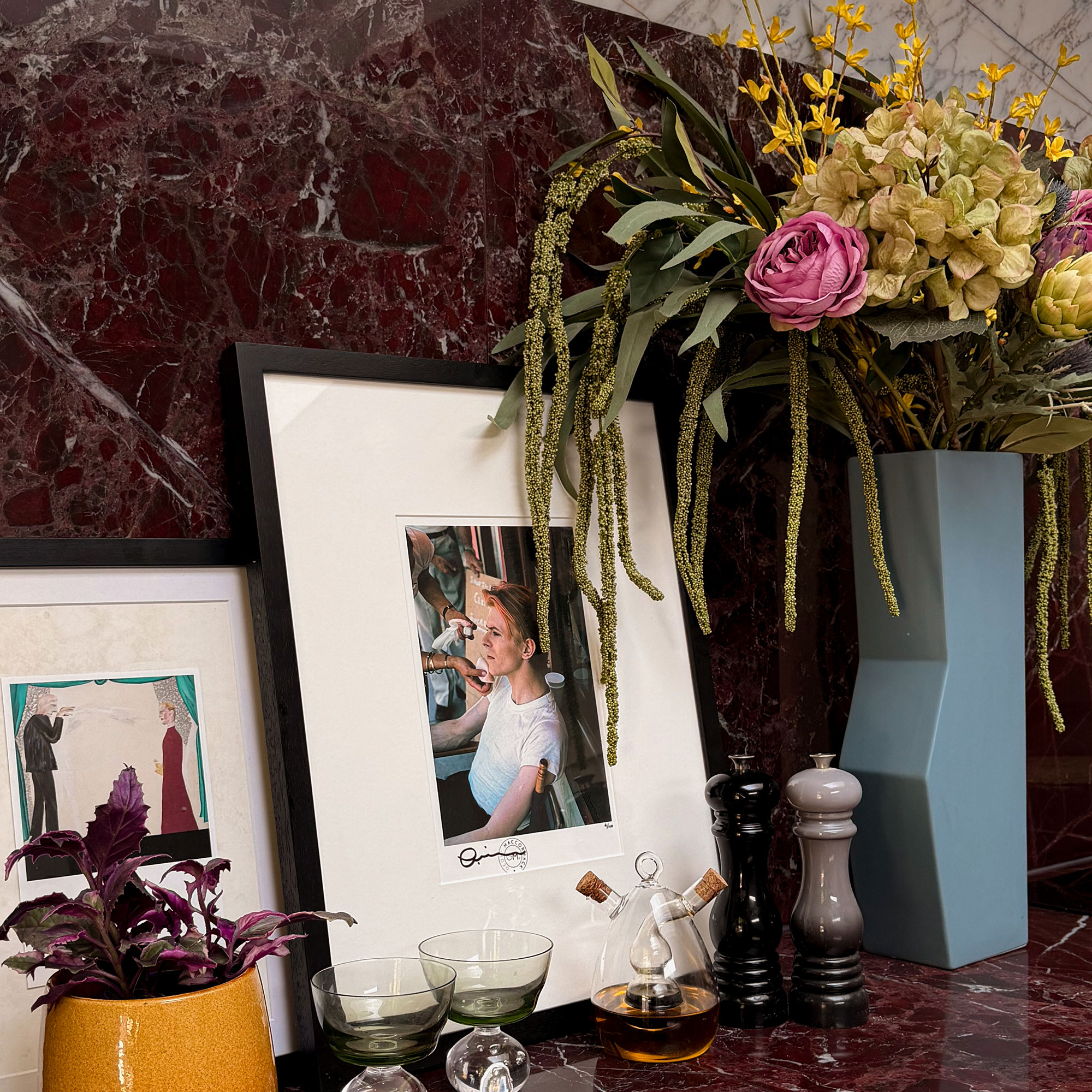 Red marble kitchen countertop and matching splashback displaying photographs, vase of flowers, salt and pepper grinders and oil dispense