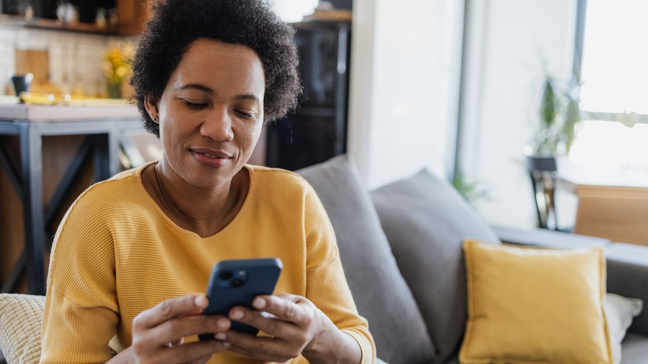 A woman smiles as she looks at her smartphone while sitting on her sofa.