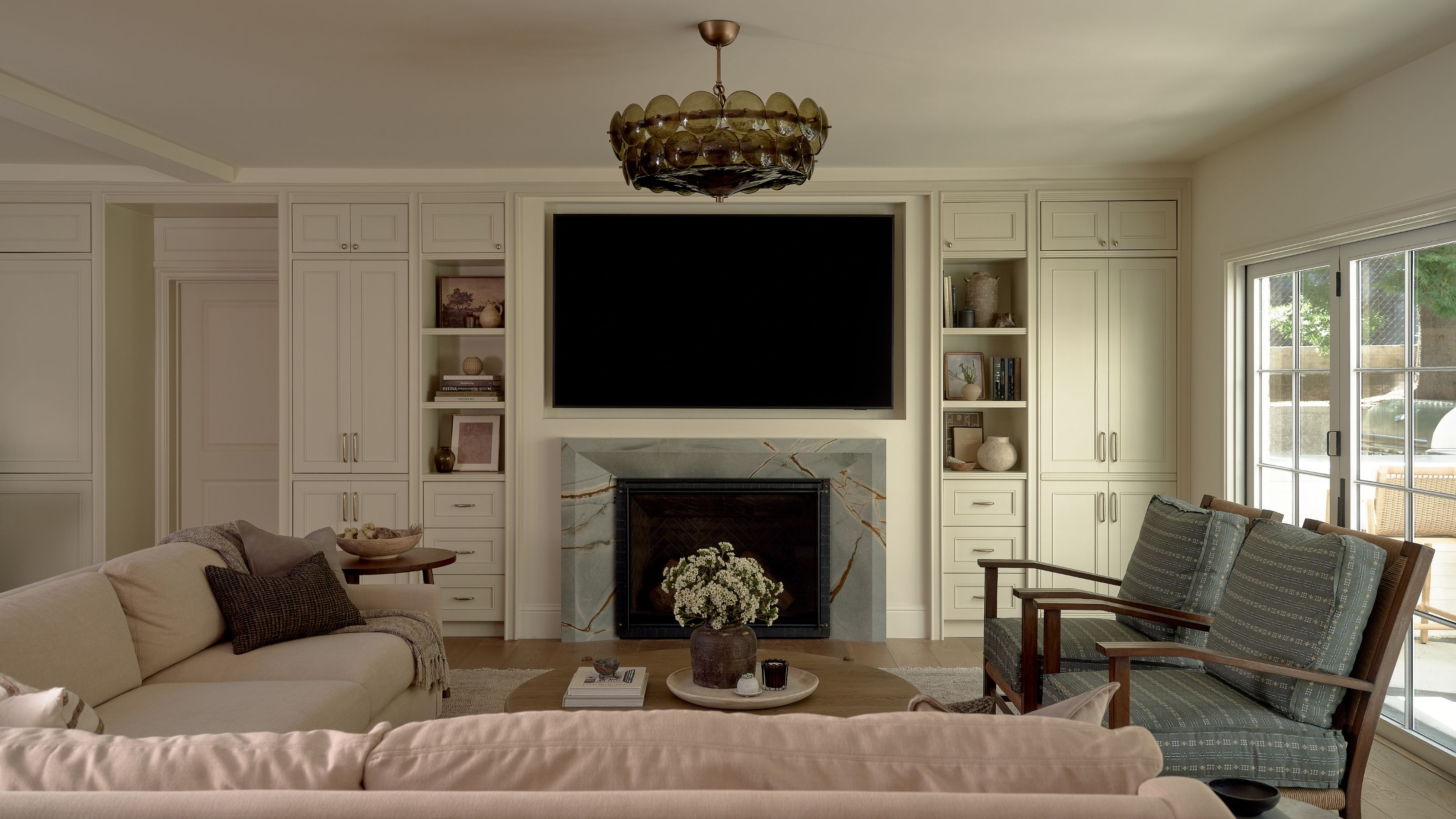 a neutral open plan sitting area with built in cabinetry, a large TV, marble fireplace, sectional couch and armchairs set in front of french doors out to the patio