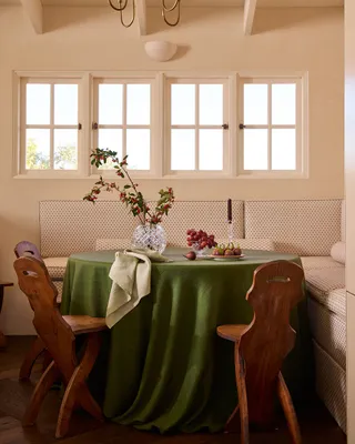 A dining table is covered in a green table cloth and decorated with wooden chairs, flowers, and fruit.