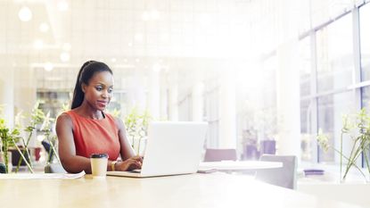 A woman sits are her desk in a corporate office and looks at her laptop, smiling.