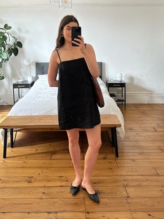 Woman in bedroom wears black mini dress, black mules