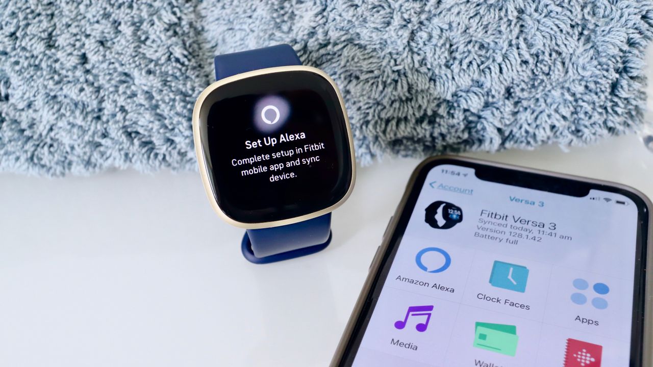 Et bl&aring;t Fitbit Versa 3 ligger ved siden af et t&aelig;ppe og en mobiltelefon.
