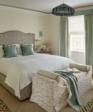 neutral bedroom with wooden floor, a patterned rug, upholstered wavy headboard and green accents with cushions and curtains