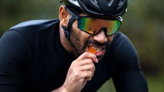 Male cyclist wearing helmet and sunglasses takes a bite out of an orange chew while riding