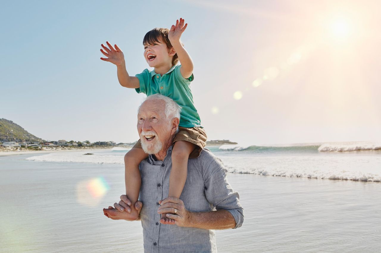 A grandfather has his grandson on his shoulders. They are at the beach.