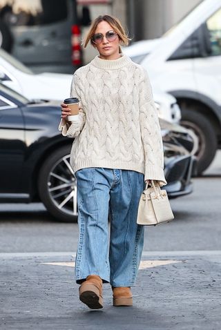 Jennifer lopez wears a white cable knit sweater, blue jeans, platform ugg boots, and a white hermes bag.