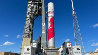 a white and red rocket stands next to its gray launch tower under a blue sky.