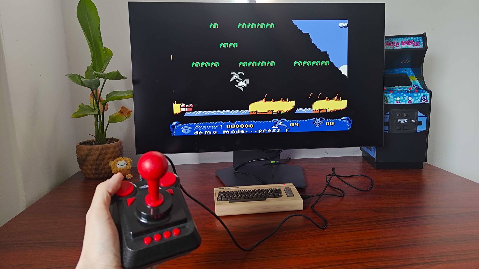 Hand holding C64 Mini joystick with C64 Mini in backdrop connected to monitor displaying Nobby the Aardvark gameplay on screen.