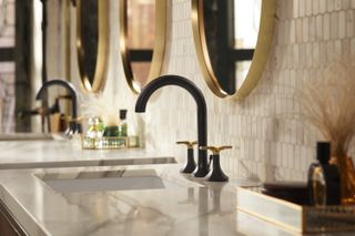 Close up image of the ‘Occasion’ faucet collection, white marble sink surround, black and gold taps, round gold framed wall mirrors, white wall tiles, baskets of toiletries on each sink