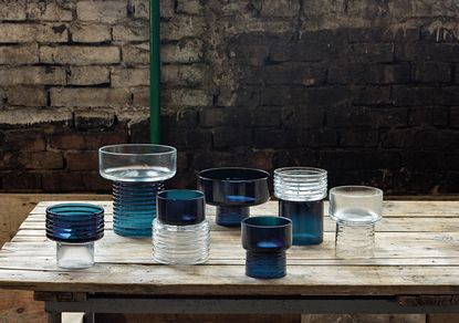 Seven cylindrical glass vessels in a mixture of blue and clear colours displayed on a table. 