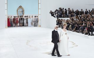 Catwalk stage, white floor with gold motif, male designer dressed in black suit, his pregnant wife in a white wedding gown, female models from the show gathered at the stage entrance, audience sat in seats watching the stage, white wall background