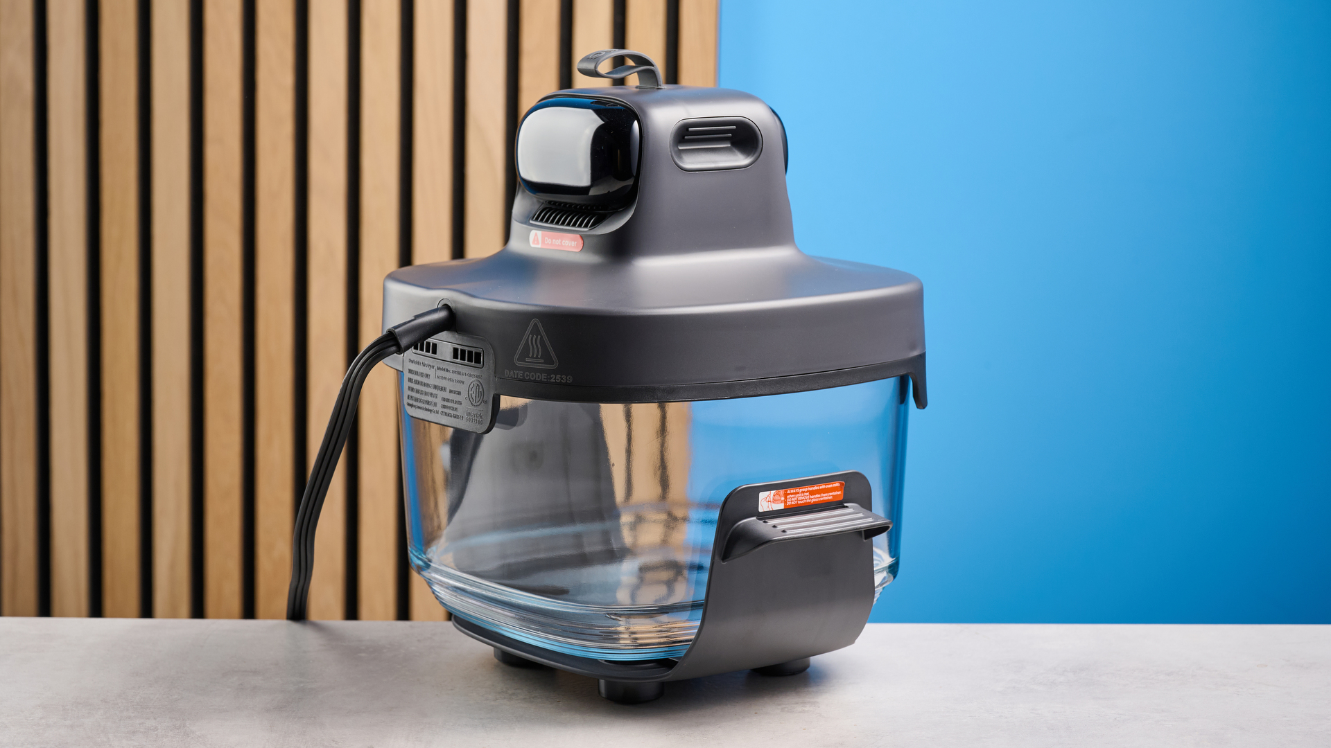 the kismile roasti air fryer with a glass cooking container photographed against a blue and wood background, showing the heating element lid