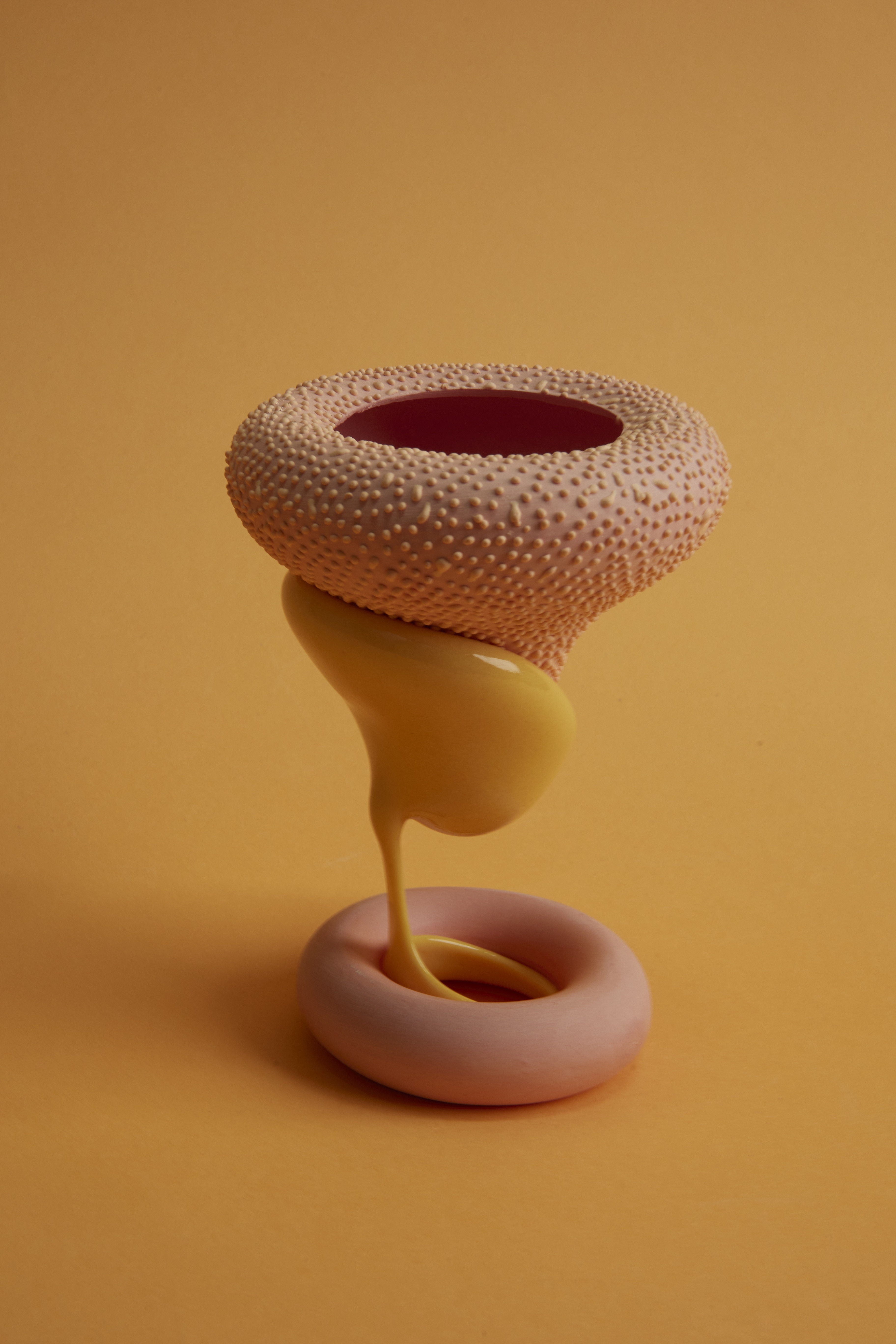 A surreal, textured ceramic sculpture in nude pink with a cylindrical shape, a dotted, top section, a liquid-like, ochre one, and a doughnut-shaped bottom sits against an ochre background.