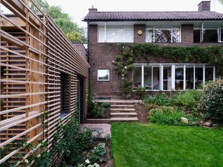 Daytime exterior view of a brick and timber house extension, brick house, green lawn, stone steps, flowers and shrubs, black frame patio window, black drainpipe, white framed glazed door, window, tall trees in the backdrop, pale sky