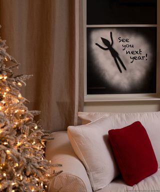 A living room with lit Christmas tree and elf on the shelf window decor with elf silhouette created with artificial snow spray paint in white