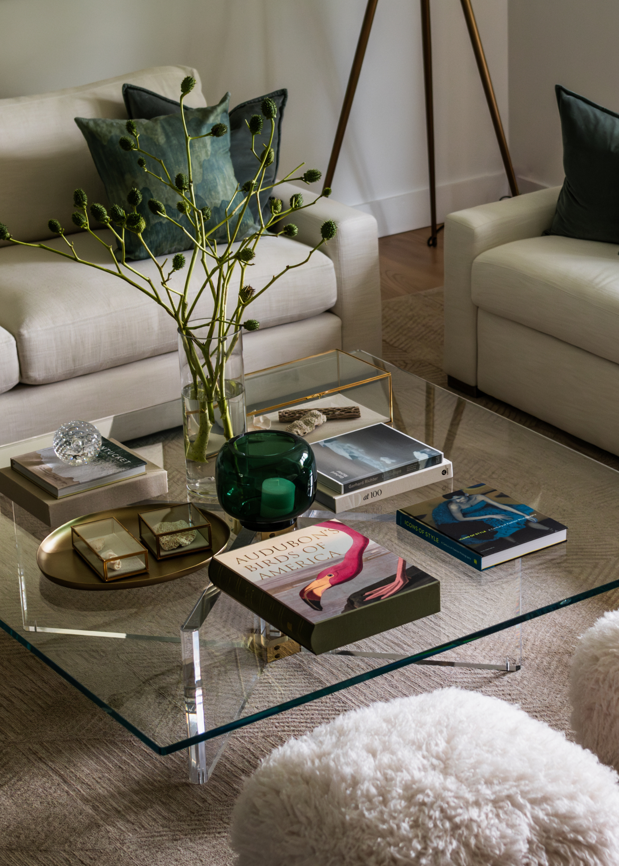 A living room with a glass coffee table with books, a candle, a vase of stems, a tray, and fuzzy pouffes