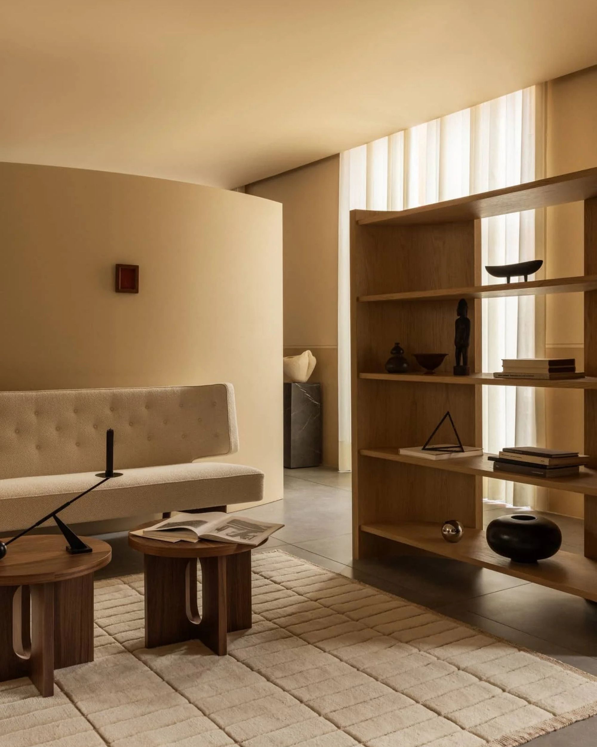 Image of large wooden shelving in a beige room with curved walls. There is a white sofa, two wooden coffee tables, and a white textured rug in the room.