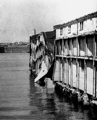 Black and white photo showing side view of dilapidated pier building,