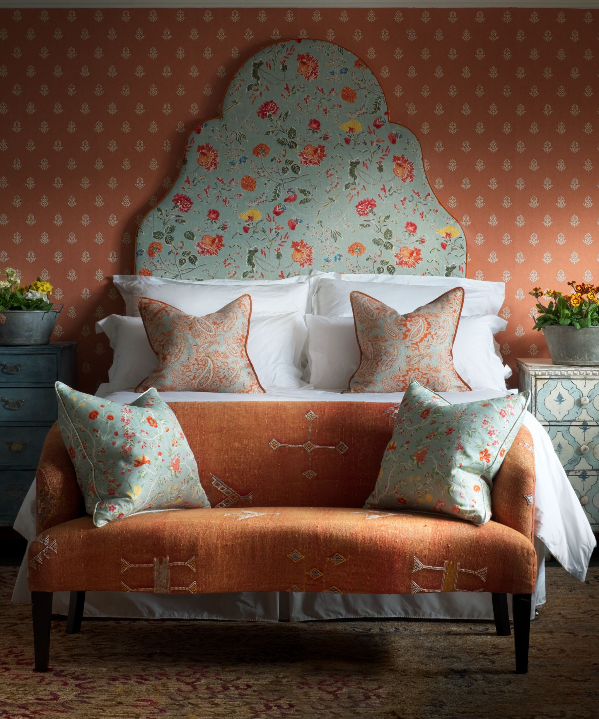 rust orange block print wallpapered bedroom with a light blue and orange supersized floral headboard, white bed linen, a rustic upholstered bench at the foot of the bed