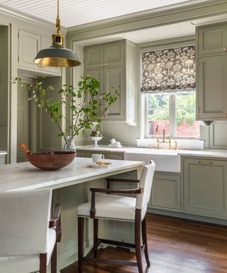 Traditional kitchen with dark wood flooring, white paneled ceiling, gray-beige painted kitchen cabinets, kitchen island with seating, patterned roman blind above sink