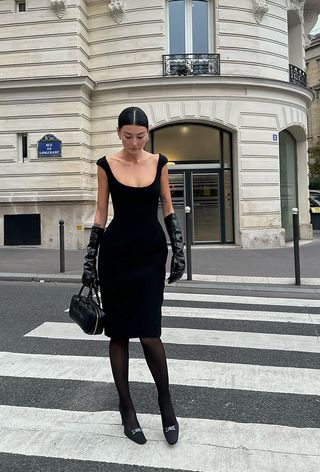 A woman wearing a black cocktail dress with black tights, black pumps, black leather gloves, and a black handbag.