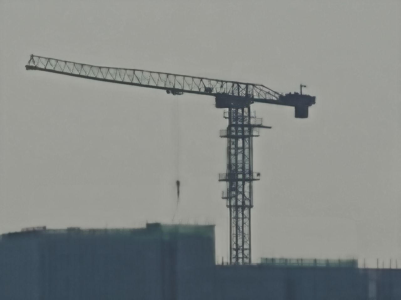 An extreme digital zoom focusing on a construction crane atop a building. The crane&rsquo;s skeletal metal arm and vertical tower are silhouetted against a flat, gray sky. The details are softened and pixelated, giving the structural lines a blurred, high-contrast appearance.