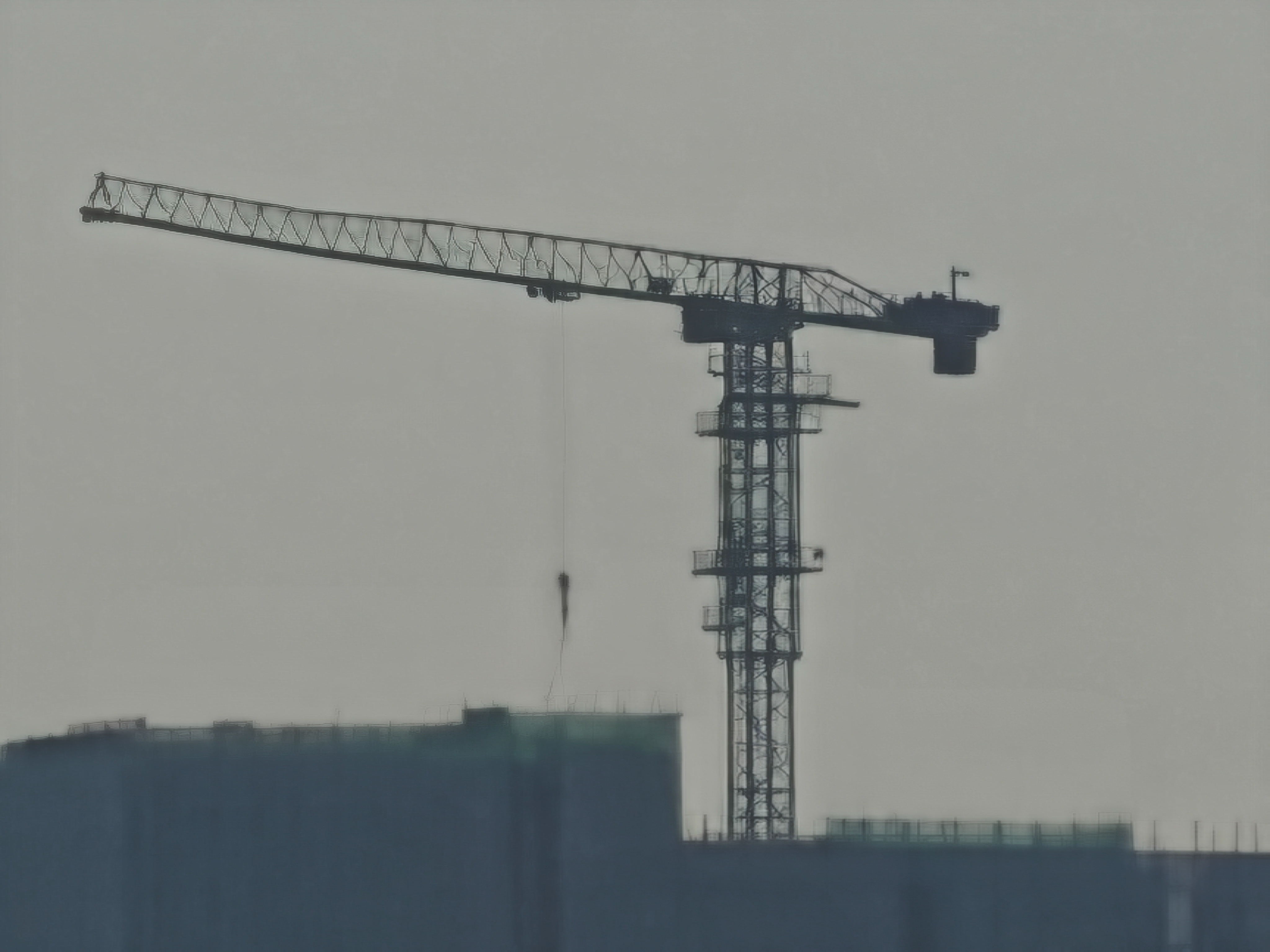 An extreme digital zoom focusing on a construction crane atop a building. The crane&rsquo;s skeletal metal arm and vertical tower are silhouetted against a flat, gray sky. The details are softened and pixelated, giving the structural lines a blurred, high-contrast appearance.