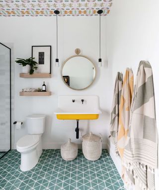 Green tiled bathroom floor, wallpapered ceiling, white walls, yellow basin, shower to left, towels hanging, mirrors, lighting,