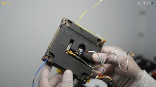 A pair of white transparent latex gloved hands holds a black square with divets at the corners, and loose wires springing from different points.