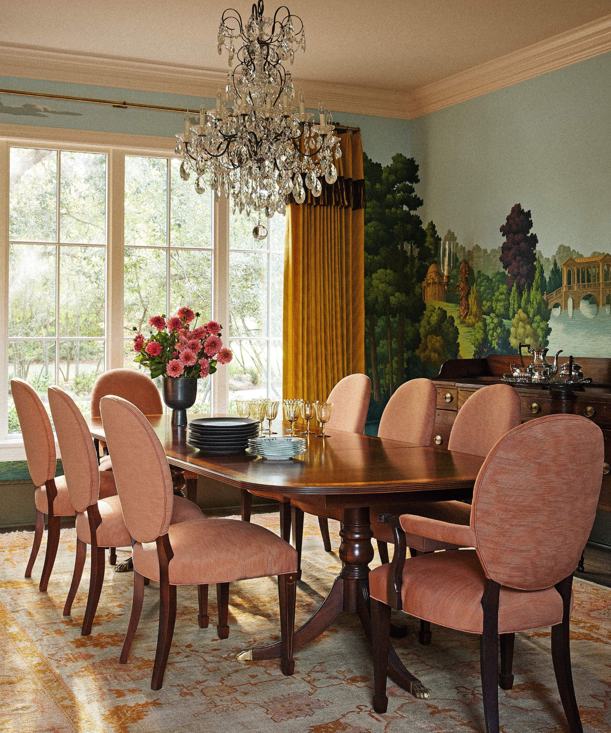 A formal dining room featuring a scenic mural wallpaper, a long mahogany table surrounded by coral-colored oval-back chairs, and a large ornate crystal chandelier