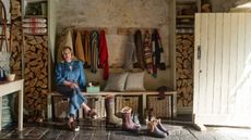 Penelope Chilvers sitting on a bench in her boot room with coat hooks behind her, woven storage baskets and a stack of logs