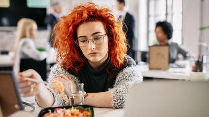 A woman in an office looks despondently at a forkful of food. There is a group of people in business attire in the background