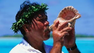 A man wearing a laurel holds a conch shell to his lips and blows into it, practicing the ancient ritual of shankh breathing