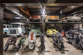 An inside park area featuring a front row of motor bikes / mopeds and a back area consisting of old sports cars including old VW cars and vans.