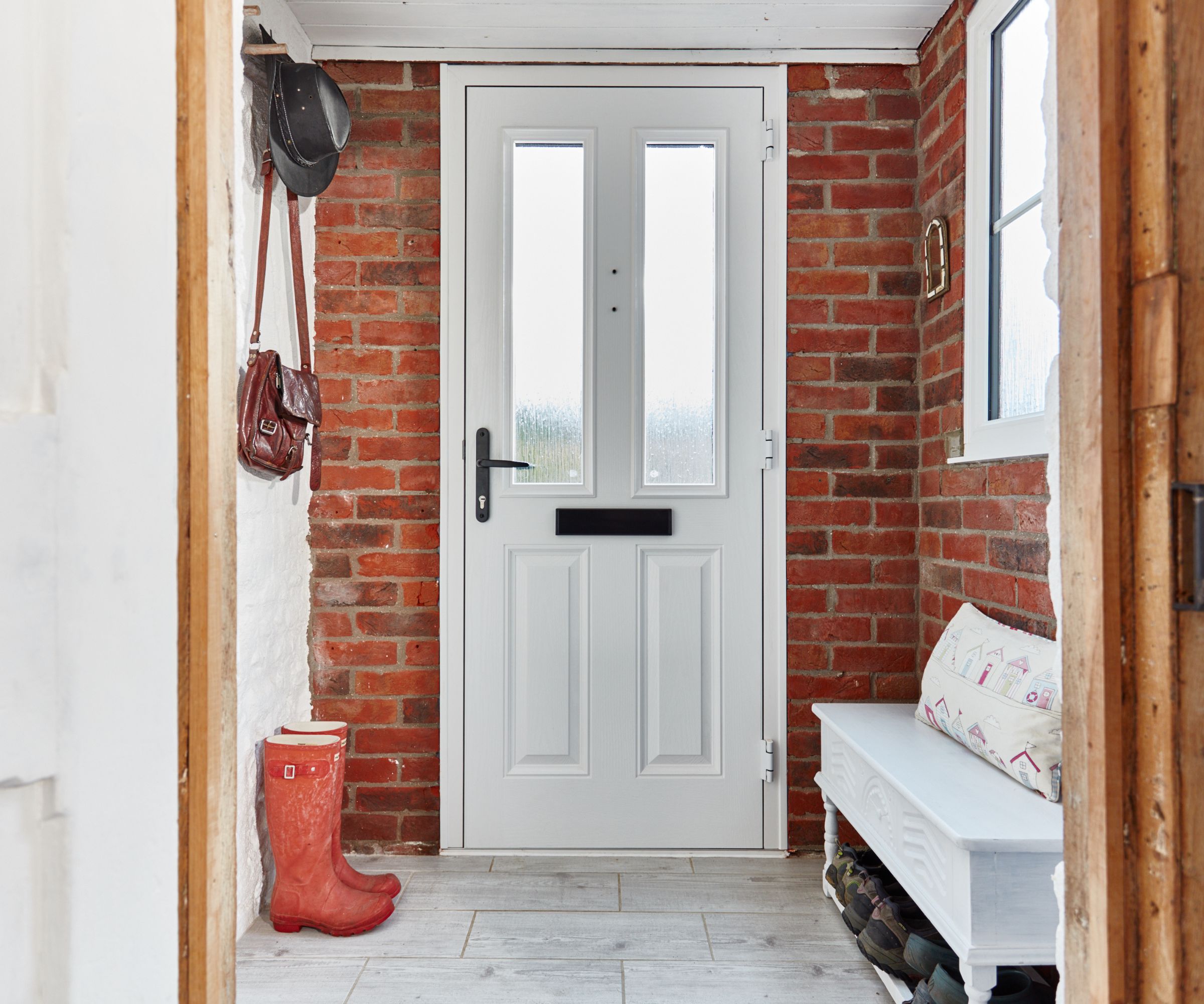 A small porch with red brick walls, a white front door with glass panels, and tiled flooring. A bench, boots, and coats add a cosy, practical touch.