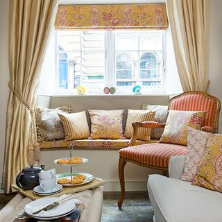 Window seat with french patterned cushions, arm chair, curtains and a table with pastries on