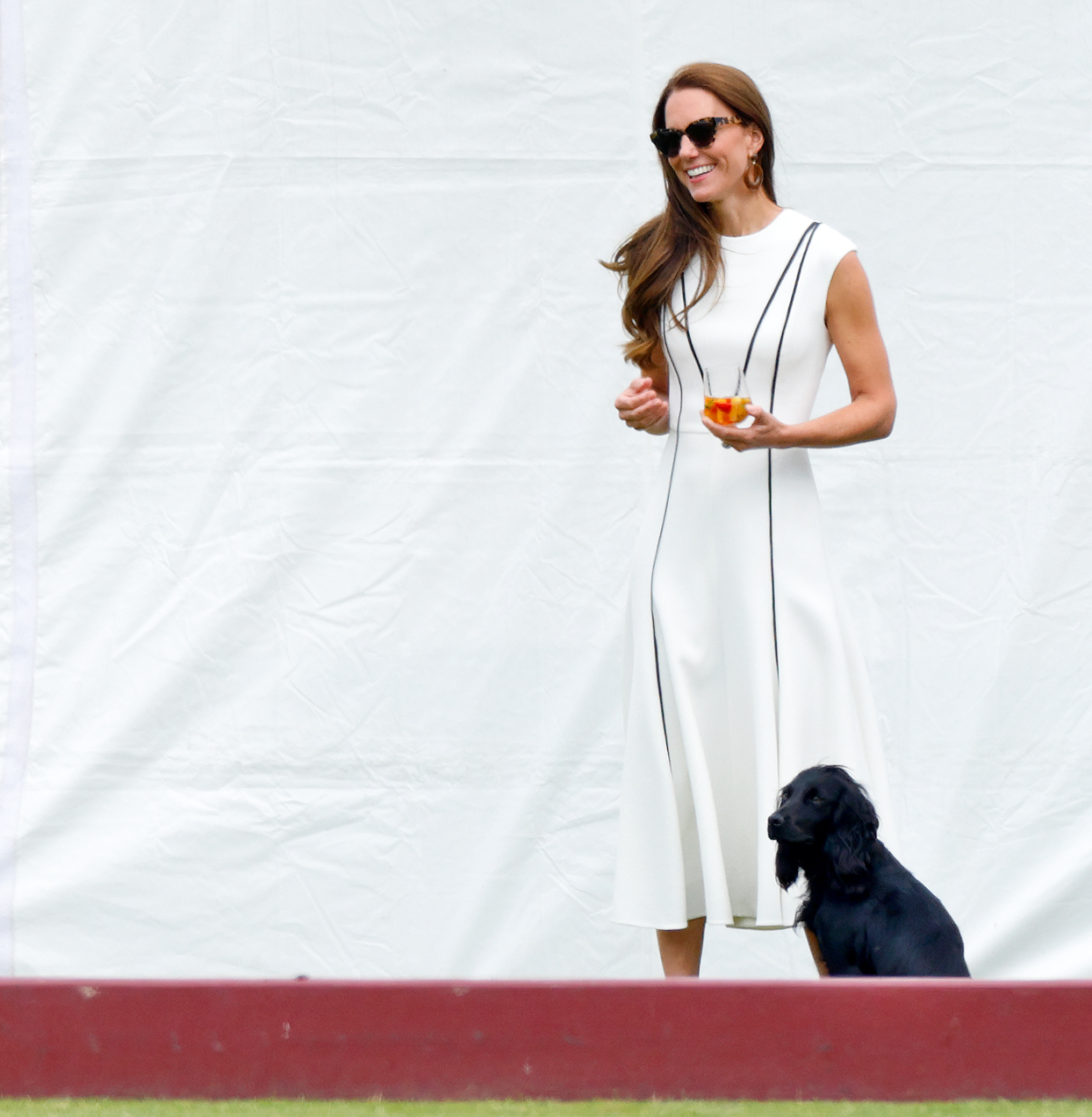 Kate Middleton wearing a white dress standing in front of her dog, Orla, and holding a glass