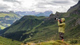 A photographer in a rain coat uses a camera with a telephoto lens surrounded by mountains