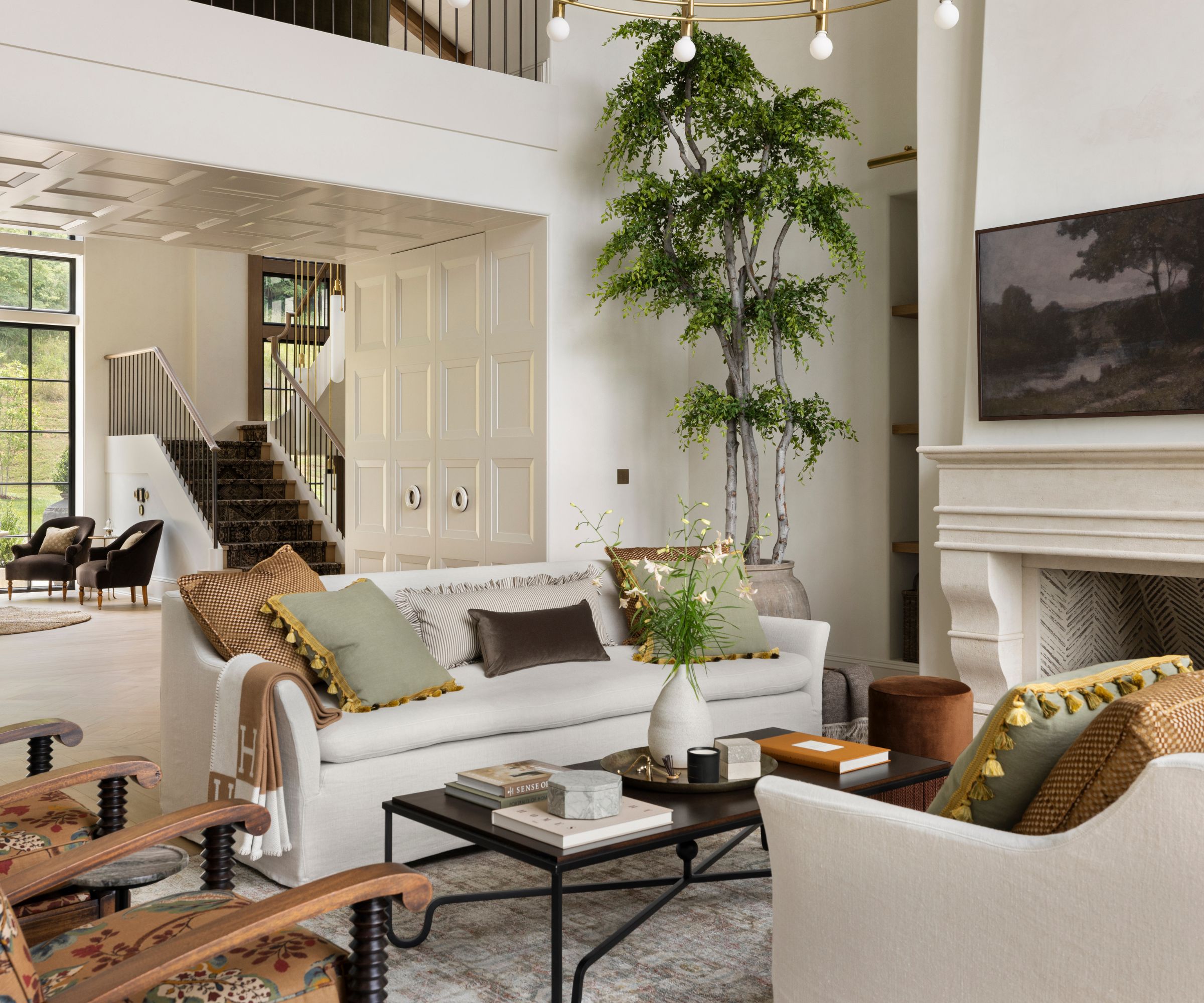 A living room with high ceilings, a large stone fireplace, two linen sofas with warm neutral cushions, a wrought-iron coffee table, and a large potted tree in the corner.