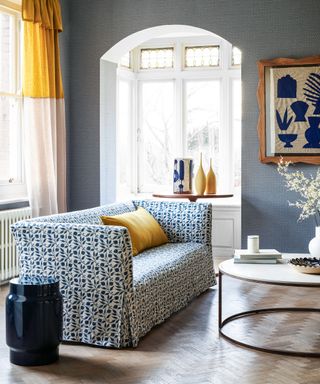 Living room with blue walls, wooden parquet flooring, blue and white upholstered floral sofa, yellow curtains and cushion.
