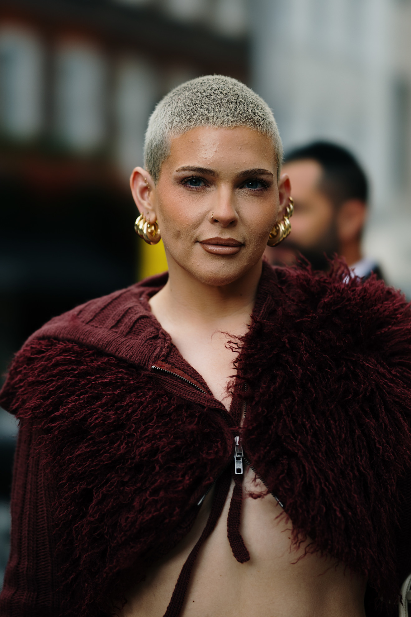 a woman with a platinum blonde buzz cut, gold hoop earrings, and a burgundy fuzzy jacket facing the camera