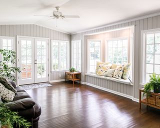 Large and open living room with windows on two sides and lots of natural light flowing in. There is a window seat on one side and a leather couch and plant on the other