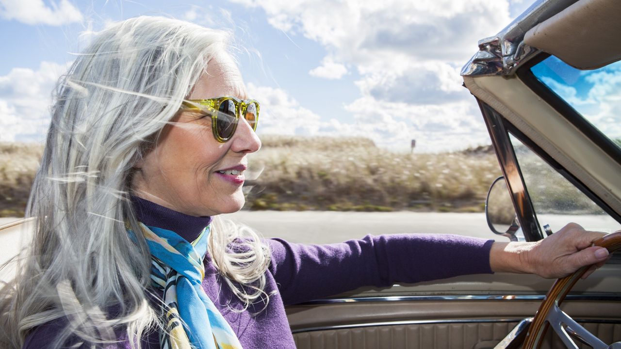 An older woman smiles as she drives a convertible with the top down.