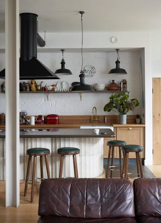 Image of a white kitchen with a textured white island that has a smooth, dark brown worktop. There are four wooden and emerald green stools, a black hutch, and wooden cabinetry. The sink has weathered brass appliances