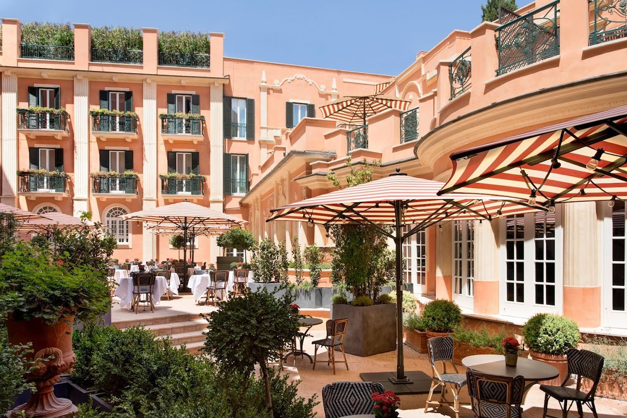 A terracotta-hued Roman terrace features stripy umbrellas, round tables, and a dense trail of plants.