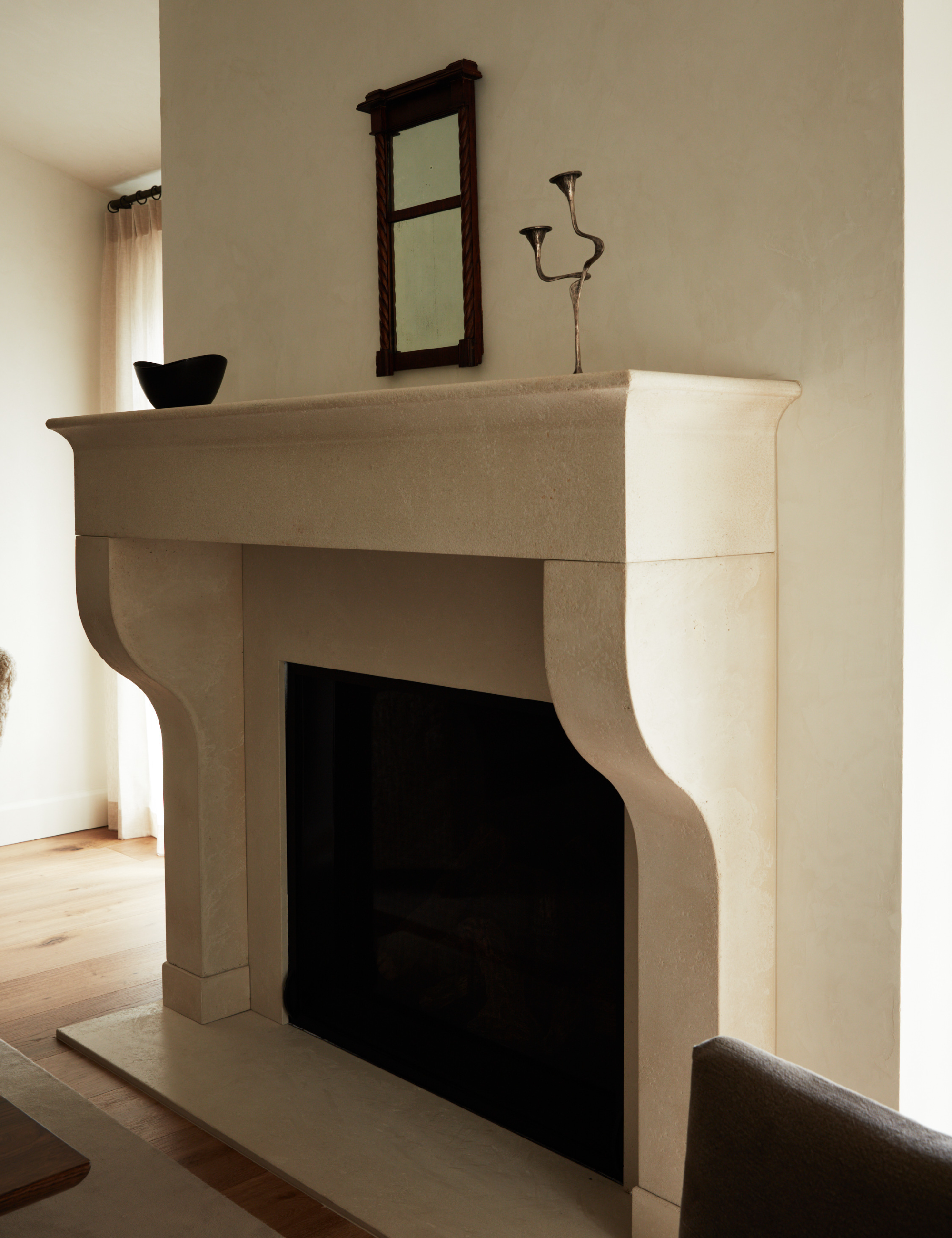 Plaster fireplace area featuring a small-scale antique wooden mirror, asymmetrical brass candelabra, and black ceramic vessel.