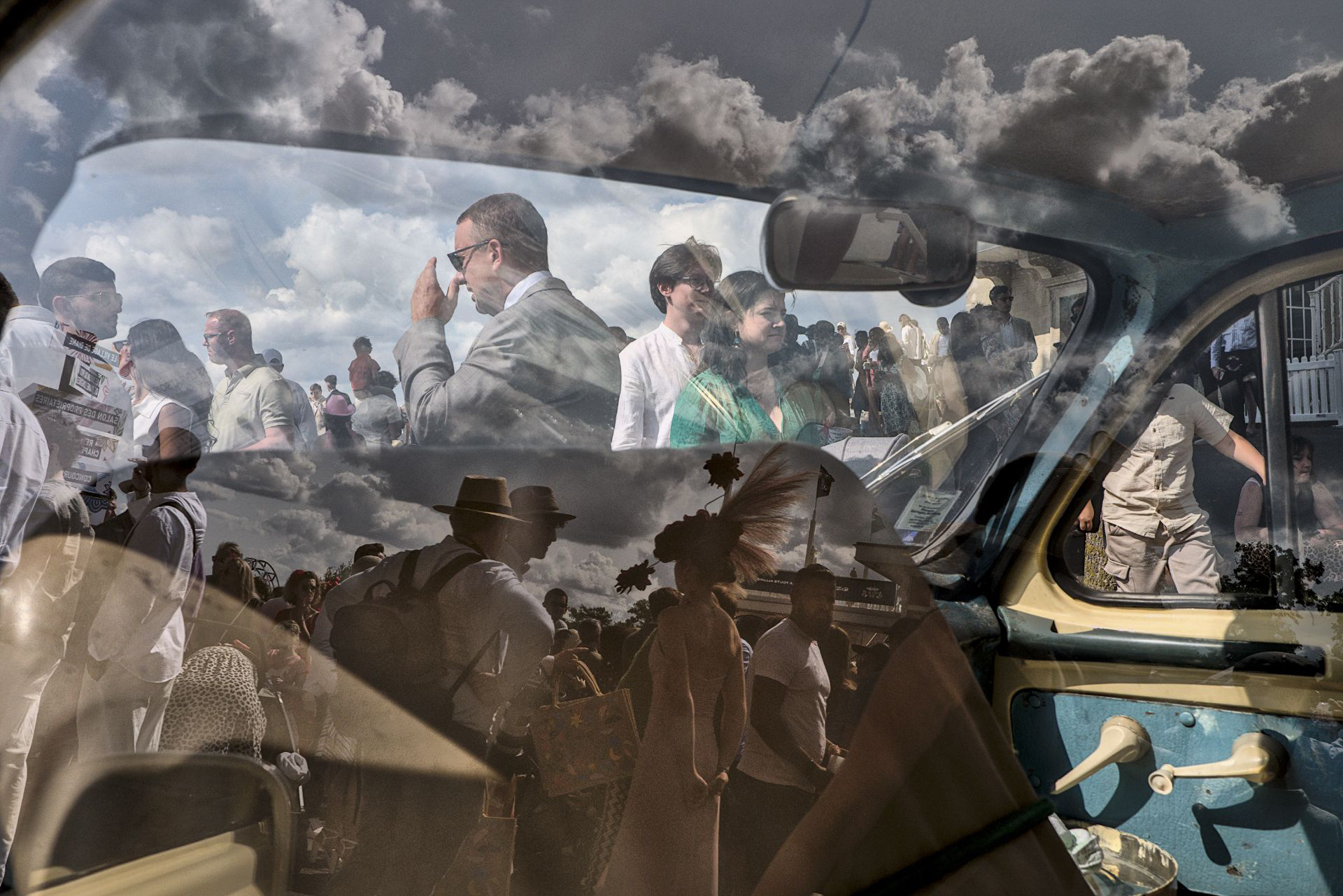 Reflection of a crowd in a car window, blending people dressed in various styles with cloudy sky and car interior details