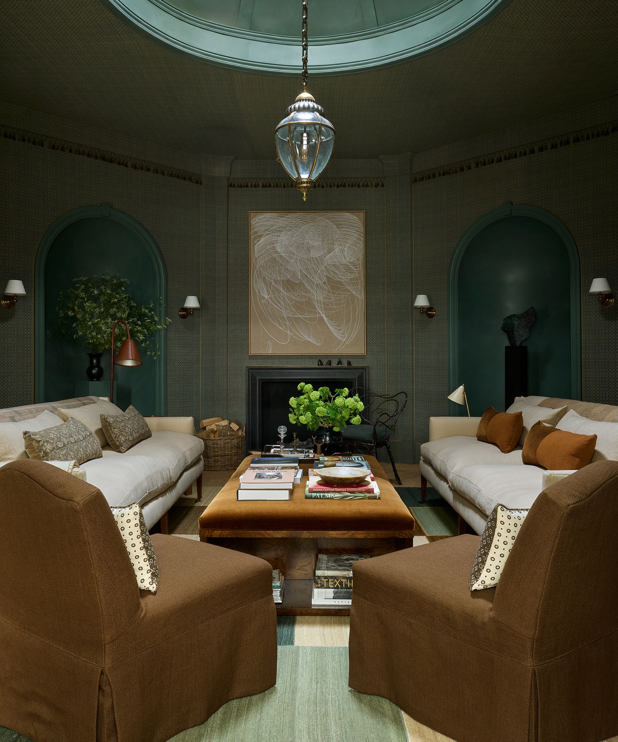 Living room with sage green grasscloth walls, alcoves with pedestals and statues, cream linen sofa, brown velvet coffee table with books on top, and deep brown slipcovered armchairs