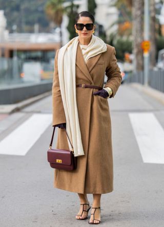 Gili Biegun wears Herskind brown wool fitted long coat with scarf, Leather gloves Léo et Violette burgundy bag, Khaite strappy sandals, Celine sunglasses, Anna Rossi earings and bracelets on November 15, 2024 in Barcelona, Spain.